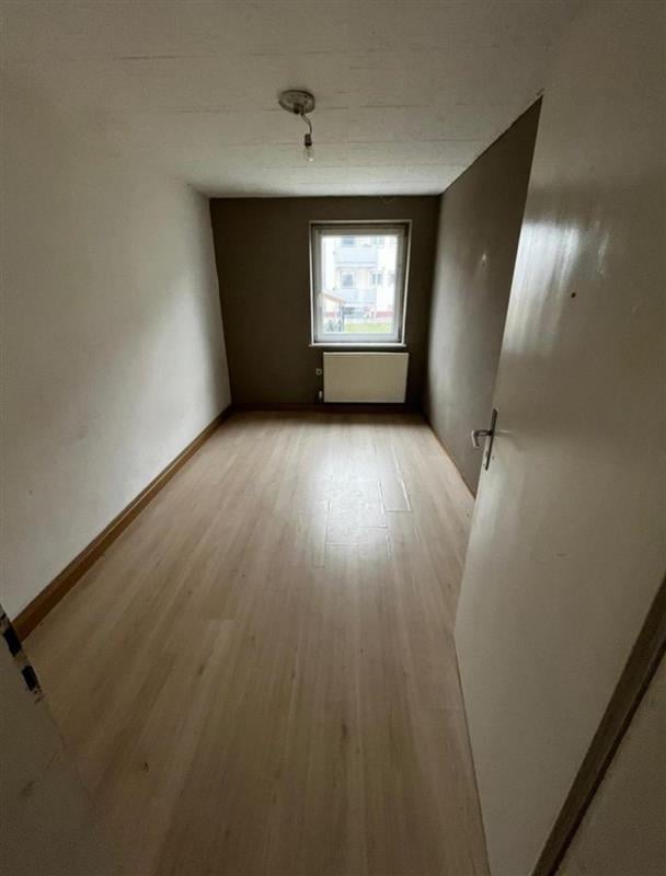 Empty apartment room with light wood flooring, white walls, window, and open door on the right side