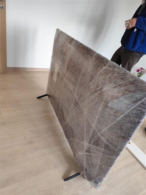 Person in blue jacket holding up a large gray upholstered panel or fabric sample in a minimalist room with wooden floor