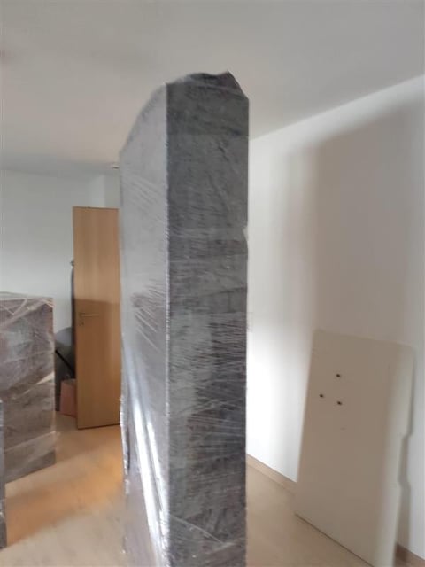 Tall dark gray wrapped object standing vertically indoors, surrounded by white and wooden materials on wooden floor