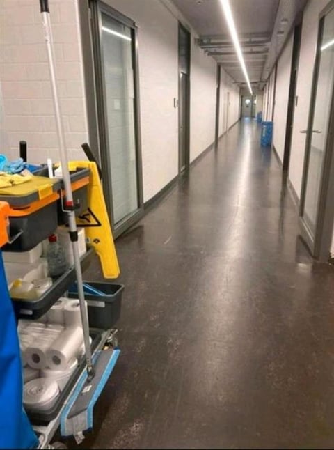 Yellow and blue cleaning cart with supplies parked in a long, empty fluorescent-lit commercial hallway with glass-fronted offices