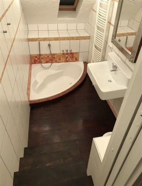 A small bathroom with white subway tile walls, dark flooring, a corner bathtub with wooden trim, pedestal sink, towel rack, and chrome mirror