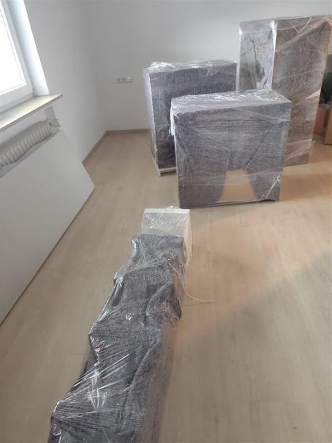 Furniture wrapped in plastic sheets stacked in an empty room with wooden flooring and a window