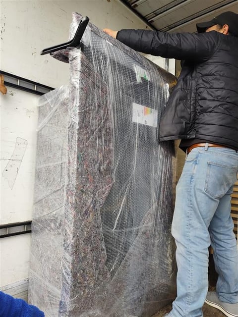 Person in black jacket and jeans unwrapping a large item covered in plastic wrap and protective bubble packaging inside a warehouse